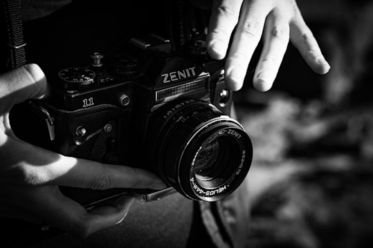 A black and white photograph features a person holding a vintage Zenit 11 camera, with both hands visible. The focus is on the camera and the hands, capturing the details of the camera&rsquo;s lens and dials. The lighting creates stark contrasts and highlights the texture of the camera and the person's skin.