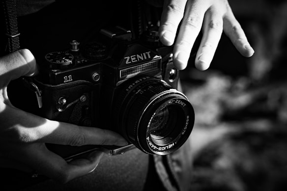 A black and white photograph features a person holding a vintage Zenit 11 camera, with both hands visible. The focus is on the camera and the hands, capturing the details of the camera&rsquo;s lens and dials. The lighting creates stark contrasts and highlights the texture of the camera and the person's skin.