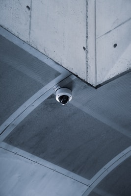 A security camera is mounted on the corner of a concrete ceiling. The ceiling appears to have a grid-like metal mesh and industrial design elements.