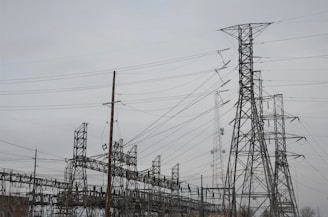 A network of high-voltage electricity pylons and transmission towers connected by numerous power lines. The structures are predominantly metallic with a lattice design, set against an overcast sky.