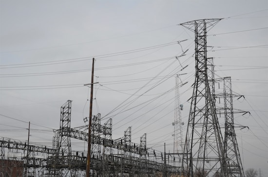 A network of high-voltage electricity pylons and transmission towers connected by numerous power lines. The structures are predominantly metallic with a lattice design, set against an overcast sky.