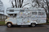 A vintage camper van is heavily covered in a variety of stickers and decals. It appears to be parked on a quiet street with trees in the background. The van shows signs of rust and wear, particularly around the wheels and edges.