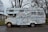 A vintage camper van is heavily covered in a variety of stickers and decals. It appears to be parked on a quiet street with trees in the background. The van shows signs of rust and wear, particularly around the wheels and edges.