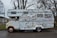 A vintage camper van is heavily covered in a variety of stickers and decals. It appears to be parked on a quiet street with trees in the background. The van shows signs of rust and wear, particularly around the wheels and edges.