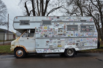 A vintage camper van is heavily covered in a variety of stickers and decals. It appears to be parked on a quiet street with trees in the background. The van shows signs of rust and wear, particularly around the wheels and edges.