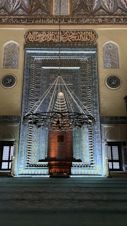 An intricately designed interior of a mosque features a prominent mihrab, beautifully adorned with geometric and floral patterns. The ambiance is illuminated by a large, ornate chandelier hanging in front of the mihrab. Arabic calligraphy is elegantly inscribed above the mihrab and along the walls, adding a spiritual atmosphere to the space.