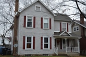 A two-story house with gray wooden siding and red shutters stands surrounded by leafless trees. The chimney is made of brick, and a satellite dish is attached to the side. The porch features decorative white railings and a gabled roof. A fenced property with adjacent homes is visible, reflecting a suburban environment during an overcast day.