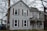 A two-story house with gray wooden siding and red shutters stands surrounded by leafless trees. The chimney is made of brick, and a satellite dish is attached to the side. The porch features decorative white railings and a gabled roof. A fenced property with adjacent homes is visible, reflecting a suburban environment during an overcast day.