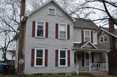 A two-story house with gray wooden siding and red shutters stands surrounded by leafless trees. The chimney is made of brick, and a satellite dish is attached to the side. The porch features decorative white railings and a gabled roof. A fenced property with adjacent homes is visible, reflecting a suburban environment during an overcast day.