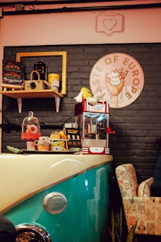 A cozy cafe scene with a vintage theme, featuring a retro popcorn machine, stuffed toys, and decorative items on a counter that resembles the front of a classic car. There's a blackboard menu, a shelf with a vintage camera, and colorful signs on a gray brick wall. Comfortable seating completes the setting.