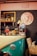 A cozy cafe scene with a vintage theme, featuring a retro popcorn machine, stuffed toys, and decorative items on a counter that resembles the front of a classic car. There's a blackboard menu, a shelf with a vintage camera, and colorful signs on a gray brick wall. Comfortable seating completes the setting.
