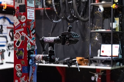 A workshop space with various tools and items. A red pole covered in colorful stickers is prominently in the foreground. Bicycle repair equipment, including a bike clamp, is attached to the pole. In the background, bicycle wheels hang from above, and there's a cluttered shelf with tools and a laptop displaying a webpage.