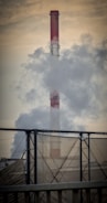 A large industrial chimney emits thick smoke into the air, with a metal structure in the foreground and a hazy sky in the background.