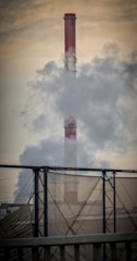 A large industrial chimney emits thick smoke into the air, with a metal structure in the foreground and a hazy sky in the background.