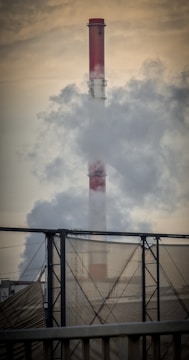 A large industrial chimney emits thick smoke into the air, with a metal structure in the foreground and a hazy sky in the background.