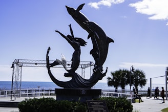 A metal sculpture depicting a mermaid reaching up toward two dolphins with the ocean in the background. The scene is set in a coastal area with palm trees and a metal framework structure visible. People can be seen walking in the distance.