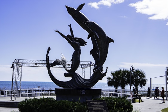 A metal sculpture depicting a mermaid reaching up toward two dolphins with the ocean in the background. The scene is set in a coastal area with palm trees and a metal framework structure visible. People can be seen walking in the distance.