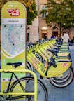 A row of bright green rental bicycles are lined up next to a map station in an urban area, likely used for bike-sharing. The station has a map and information for commuters. In the background, there are blurred figures of people and buildings with trees indicating a park-like setting.