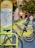 A row of bright green rental bicycles are lined up next to a map station in an urban area, likely used for bike-sharing. The station has a map and information for commuters. In the background, there are blurred figures of people and buildings with trees indicating a park-like setting.