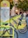 A row of bright green rental bicycles are lined up next to a map station in an urban area, likely used for bike-sharing. The station has a map and information for commuters. In the background, there are blurred figures of people and buildings with trees indicating a park-like setting.