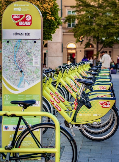 A row of bright green rental bicycles are lined up next to a map station in an urban area, likely used for bike-sharing. The station has a map and information for commuters. In the background, there are blurred figures of people and buildings with trees indicating a park-like setting.