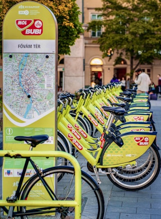 A row of bright green rental bicycles are lined up next to a map station in an urban area, likely used for bike-sharing. The station has a map and information for commuters. In the background, there are blurred figures of people and buildings with trees indicating a park-like setting.