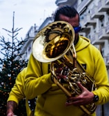 A person in a yellow hoodie plays a brass instrument, possibly a tuba or euphonium, on a city street. The reflection of buildings is visible on the shiny surface of the instrument. A decorated Christmas tree with lights stands in the background, hinting at a festive atmosphere.