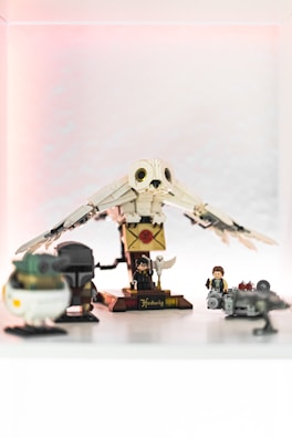 A collection of LEGO figures and objects on display, including a large owl with spread wings, a letter, and several miniature characters. The scene is set against a softly lit background with a pink hue.