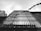 A black and white photograph of a modern building taken from a low angle, emphasizing vertical lines created by window panes and metal bars. Flags or banners are seen fluttering in the wind above the structure, creating a dynamic element against the static architecture.