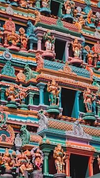 A beautifully intricate facade of a Hindu temple featuring numerous colorful sculptures depicting deities and mythical figures. The vibrant artwork is detailed with a variety of vivid colors, including teal, orange, pink, and yellow, showcasing traditional architectural elements and artistry.