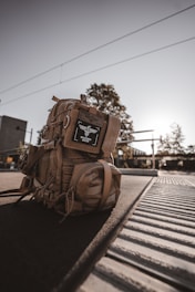 A tan tactical backpack is positioned on a train platform, illuminated by warm, backlit sunlight. The backpack features various compartments and straps, with a label displaying 'Invader Gear.' The metal tracks and grooves of the platform are visible, while trees and a modern building can be seen in the blurred background.
