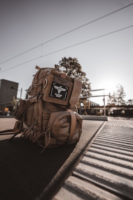 A tan tactical backpack is positioned on a train platform, illuminated by warm, backlit sunlight. The backpack features various compartments and straps, with a label displaying 'Invader Gear.' The metal tracks and grooves of the platform are visible, while trees and a modern building can be seen in the blurred background.