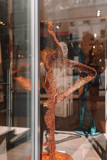 An intricate sculpture made from a network of orange metal rings is displayed inside what appears to be an art gallery. The sculpture resembles a human figure in a dynamic pose. Another similar blue sculpture can be seen in the background. The setting is indoors with well-lit surroundings and the reflections on the glass add complexity to the visual.