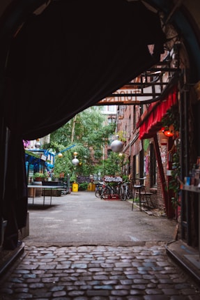 A narrow cobblestone alleyway is flanked by brick buildings adorned with various street art. There are bicycles parked against a wall, tables and chairs set along the sides, and vibrant foliage adding greenery to the scene. Decorative lights hang across the space, contributing to a cozy, eclectic vibe.