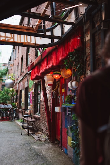 A narrow alleyway lined with brick buildings adorned with vibrant murals and colorful decorations. There are bicycles, chairs, and various hanging lights and plants contributing to a bohemian ambiance. The scene is partially enclosed by structures overhead, with a cozy and eclectic cafe or shop entrance visible.
