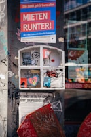 A heavily graffitied urban wall featuring various posters and stickers. The large poster at the top, containing text in German, stands out with bold colors. Below it, a closed window frame with more stickers and graffiti creates a collage effect. These elements convey messages about social issues and activism. The surrounding wall is covered with tagged graffiti, adding to the urban aesthetic.