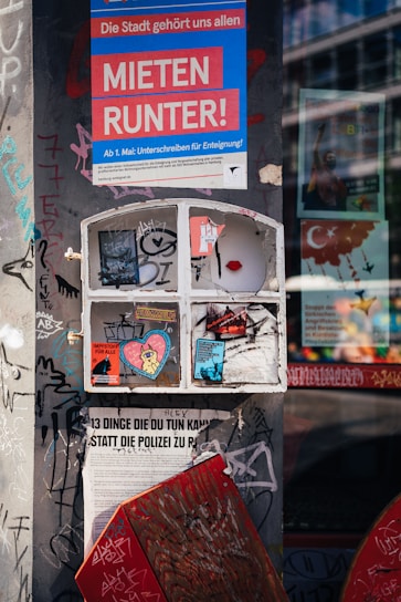 A heavily graffitied urban wall featuring various posters and stickers. The large poster at the top, containing text in German, stands out with bold colors. Below it, a closed window frame with more stickers and graffiti creates a collage effect. These elements convey messages about social issues and activism. The surrounding wall is covered with tagged graffiti, adding to the urban aesthetic.