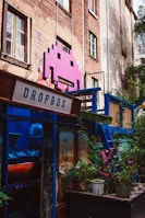 A vibrant, artistic space adorns an urban building with a large pink space invader icon mounted on the brick wall. The area is colorful, featuring a mix of greenery and various structures painted in blue. A sign labeled 'Dropbox' is present, and there is a window filled with plants and eclectic decoration, creating a lively and creative atmosphere.