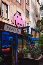 A vibrant, artistic space adorns an urban building with a large pink space invader icon mounted on the brick wall. The area is colorful, featuring a mix of greenery and various structures painted in blue. A sign labeled 'Dropbox' is present, and there is a window filled with plants and eclectic decoration, creating a lively and creative atmosphere.