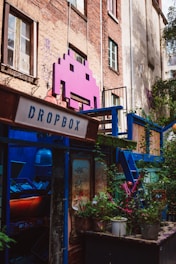A vibrant, artistic space adorns an urban building with a large pink space invader icon mounted on the brick wall. The area is colorful, featuring a mix of greenery and various structures painted in blue. A sign labeled 'Dropbox' is present, and there is a window filled with plants and eclectic decoration, creating a lively and creative atmosphere.