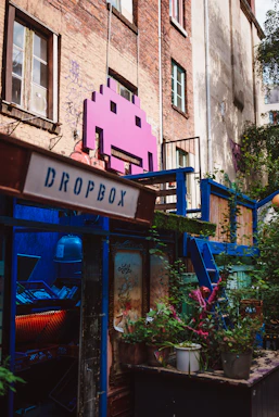 A vibrant, artistic space adorns an urban building with a large pink space invader icon mounted on the brick wall. The area is colorful, featuring a mix of greenery and various structures painted in blue. A sign labeled 'Dropbox' is present, and there is a window filled with plants and eclectic decoration, creating a lively and creative atmosphere.