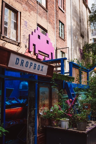 A vibrant, artistic space adorns an urban building with a large pink space invader icon mounted on the brick wall. The area is colorful, featuring a mix of greenery and various structures painted in blue. A sign labeled 'Dropbox' is present, and there is a window filled with plants and eclectic decoration, creating a lively and creative atmosphere.