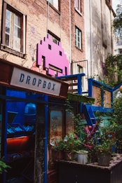 A vibrant, artistic space adorns an urban building with a large pink space invader icon mounted on the brick wall. The area is colorful, featuring a mix of greenery and various structures painted in blue. A sign labeled 'Dropbox' is present, and there is a window filled with plants and eclectic decoration, creating a lively and creative atmosphere.