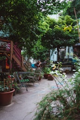 An urban garden scene featuring wooden chairs and tables scattered around a paved area. Potted plants abound, with lush greenery dominating the space. A wooden stairway leads to a higher level, and bicycles and people are visible in the background. The atmosphere suggests a cozy, communal outdoor area.