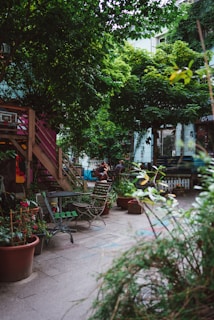 An urban garden scene featuring wooden chairs and tables scattered around a paved area. Potted plants abound, with lush greenery dominating the space. A wooden stairway leads to a higher level, and bicycles and people are visible in the background. The atmosphere suggests a cozy, communal outdoor area.