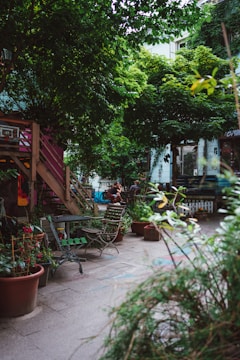 An urban garden scene featuring wooden chairs and tables scattered around a paved area. Potted plants abound, with lush greenery dominating the space. A wooden stairway leads to a higher level, and bicycles and people are visible in the background. The atmosphere suggests a cozy, communal outdoor area.