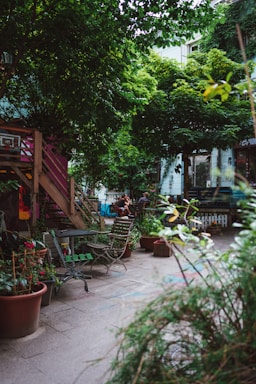 An urban garden scene featuring wooden chairs and tables scattered around a paved area. Potted plants abound, with lush greenery dominating the space. A wooden stairway leads to a higher level, and bicycles and people are visible in the background. The atmosphere suggests a cozy, communal outdoor area.