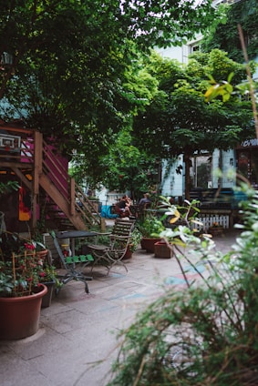 An urban garden scene featuring wooden chairs and tables scattered around a paved area. Potted plants abound, with lush greenery dominating the space. A wooden stairway leads to a higher level, and bicycles and people are visible in the background. The atmosphere suggests a cozy, communal outdoor area.