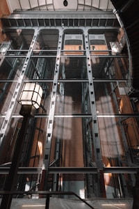 An industrial and architectural space featuring a tall, multi-level elevator shaft with metal grating and structural beams. The image has geometric shapes and lines, including a prominent lamp with a square design in the foreground.