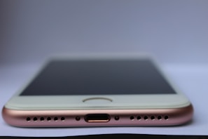 A close-up view of a smartphone lying flat on a surface with a focus on the bottom part of the device. Visible components include the charging port, speaker grills, and the edge of the screen. The phone has a metallic and sleek design.